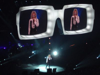 Le chanteur Polnareff en concert avec des lunettes géantentes servant d'écran au dessus de la scène