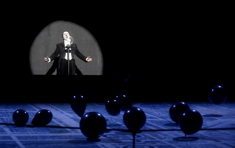 Un homme grimé en Hitler parle à un micro devant des ballons noirs au sol.